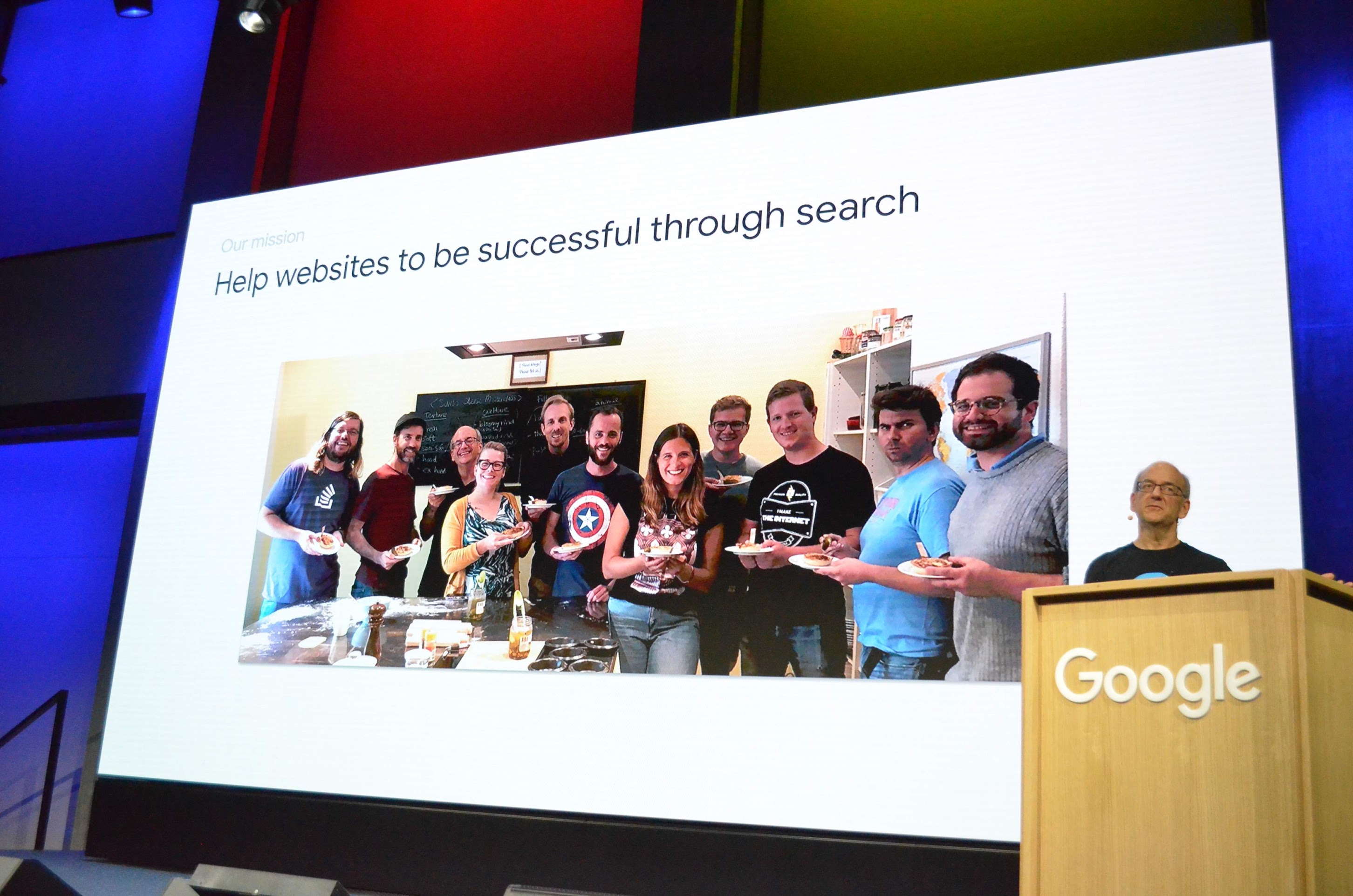 A picture of the Search Central Live Zurich event in 2019 with John Mueller in front of a slide with the title 'Help websites to be successful through Search' and a photo of the extended Search Relations team at Google.