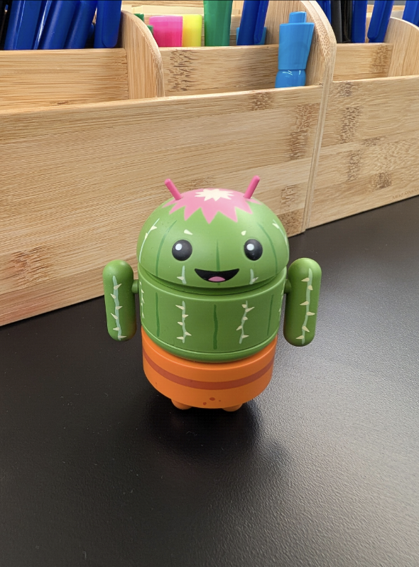 Um pequeno robô Android verde com um design parecido com um cacto está sentado em uma superfície preta.